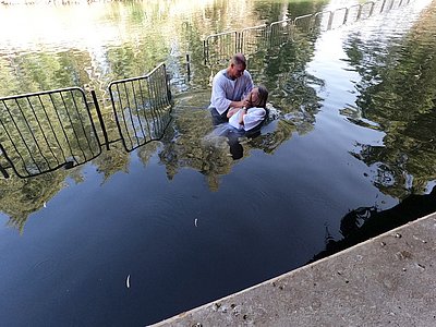 A man in a white robe baptizing a woman also in a white robe in the sea, blocked from the bigger pool of water by a gate