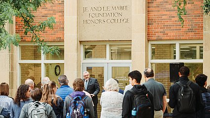 A group of people standing in front of J.E. and L.E. Mabee Foundation Honors College American Heritage building looking towards Provost Kent Gallaher as he presents a speech
