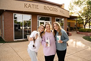 Photograph of the front of Katie Rogers Hall with three female students smiling with drinks in their hands