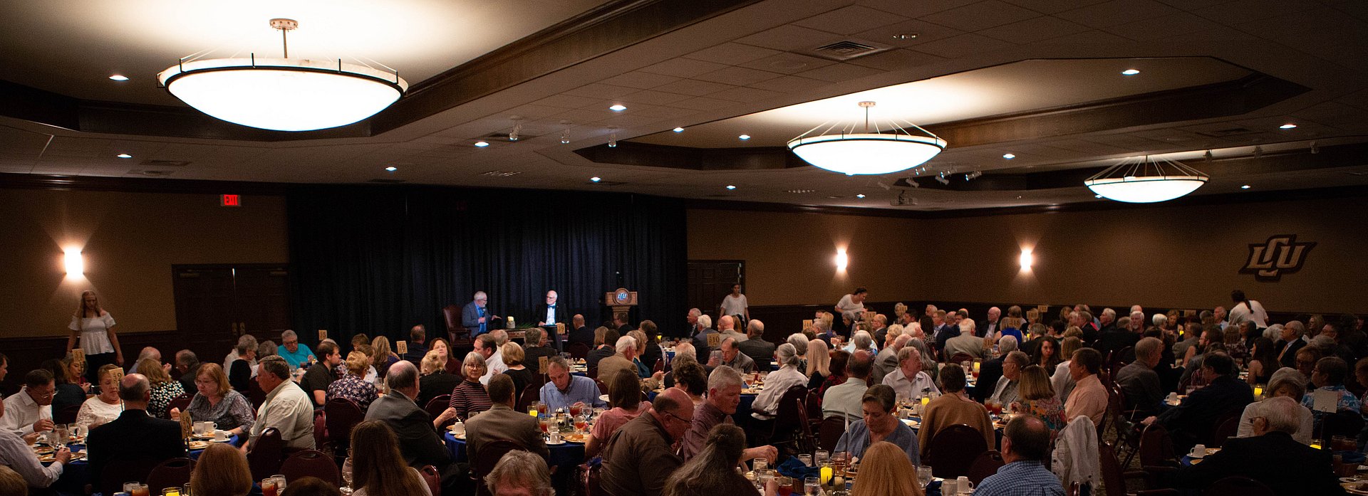 Large gathering inside the Baker Conference Center with numerous seated attendees at round tables, facing a stage with two presenters and an LCU podium to the right of them under dim overhead lighting