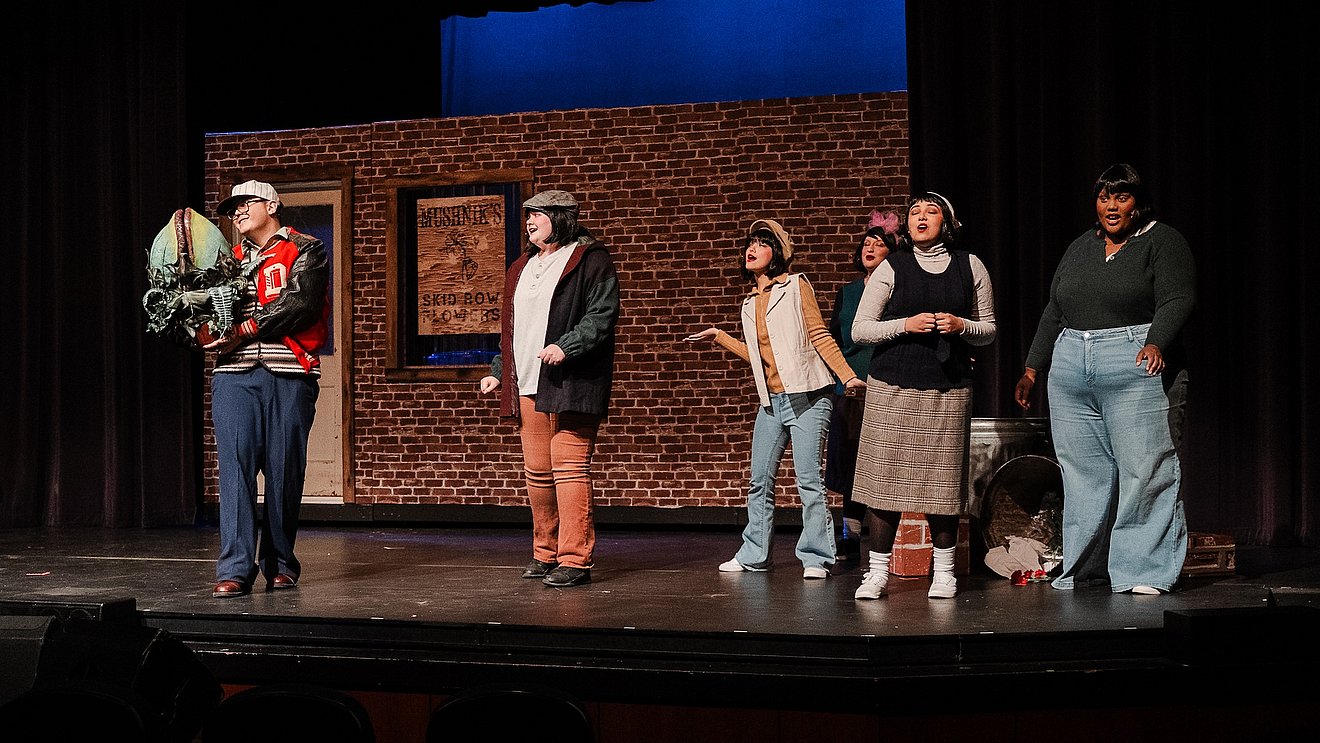 Six students on stage in the auditorium in front of a brick backdrop with a door and window, playing their part in the musical, five are off to the right side, and one man is being showcased, he is holding a Venus Flytrap puppet