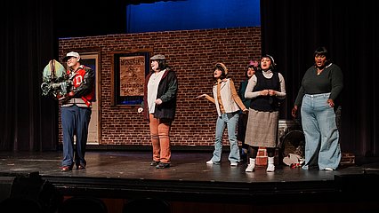 Six students on stage in the auditorium in front of a brick backdrop with a door and window, playing their part in the musical, five are off to the right side, and one man is being showcased, he is holding a Venus Flytrap puppet