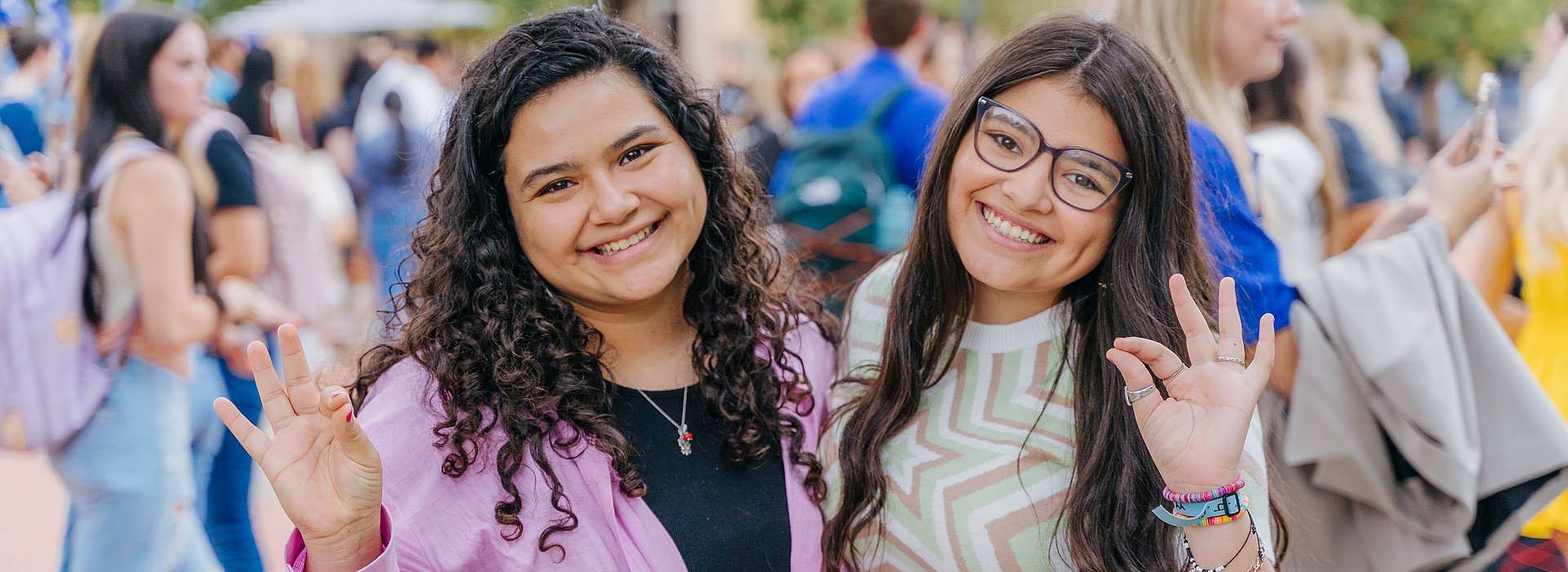Two students in front of a group of students walking, smiling at the camera and holding up the Chap hand signal