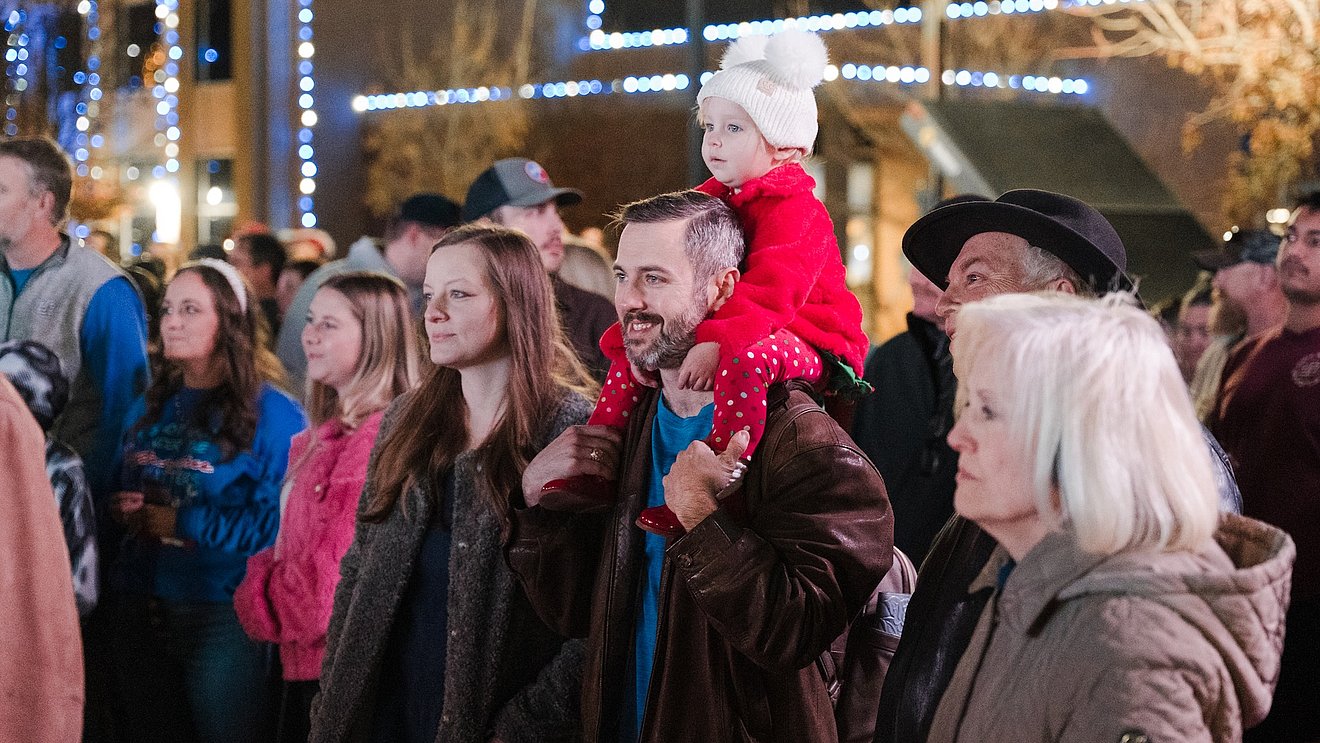 A crowd of cheerful onlookers stand enjoying the festivities in front of buildings with Christmas lights