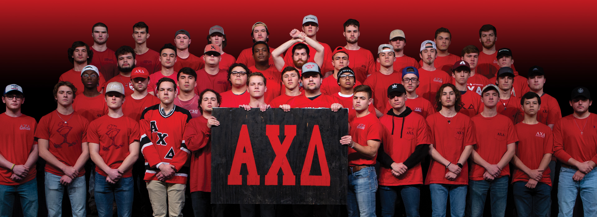 A group of men in Alpha Chi Delta wearing red shirts standing on risers and facing towards the camera with straight faces, in the front row, you see hands down and crossed, in the middle of the group, a man is throwing the Alpha Chi symbol and four men in the front middle are holding a black sign with their Greek Letters in red