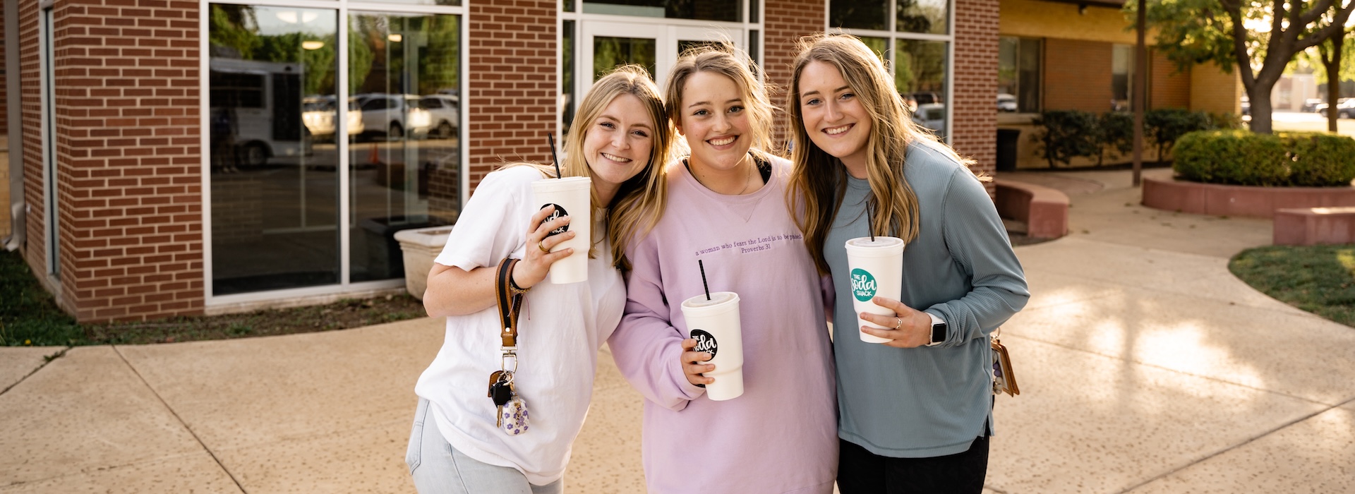 Photograph of three smiling female students in front of Katie Rogers Hall with drinks in their hands.